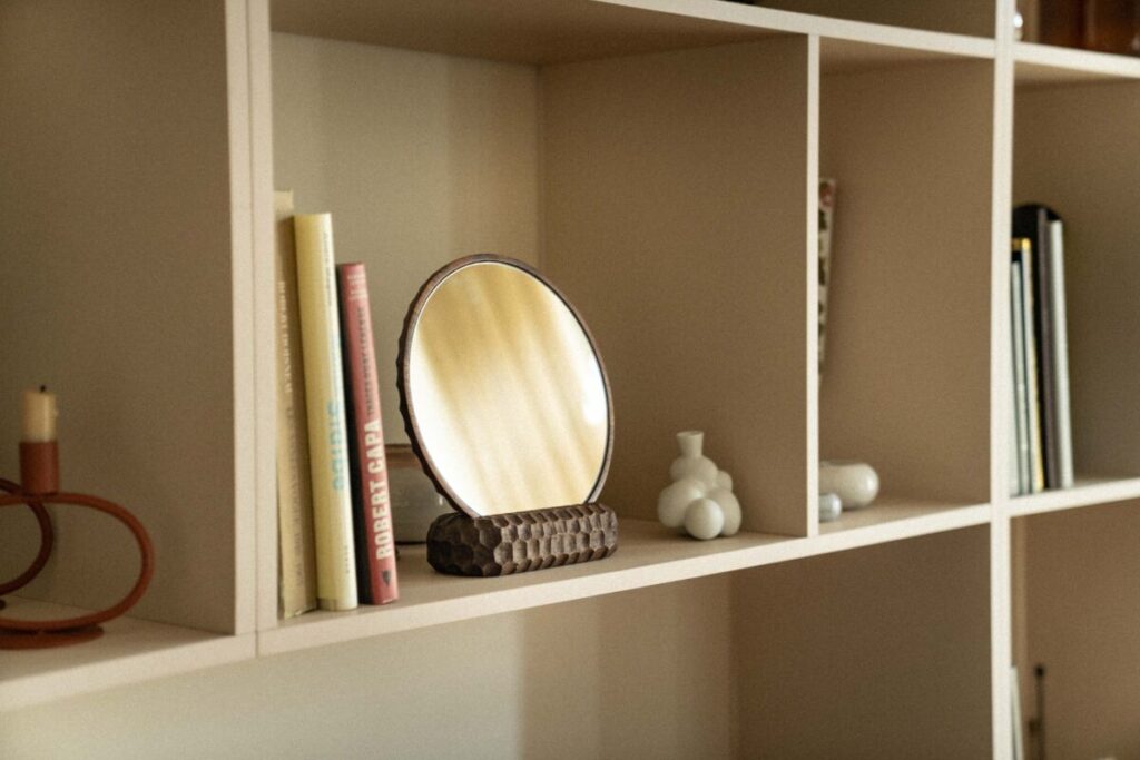 WAGA Garda Sculptural Table Mirror — ručno rezbарено ogledalo od tamnog orahovog drveta s teksturiranom bazom, postavljeno na polici između knjiga i keramičkih detalja u toplom, neutralnom interijeru