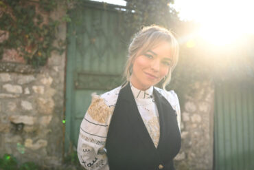 Alma Subašić-Omerović stoji ispred kamenog zida i zelenih vrata, u zlatnom svjetlu zalaska sunca, u crnom prsluku i blistavoj vezanoj bluzi