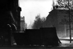 Crno-bijela fotografija puste i maglovite sarajevske ulice pod snijegom, gdje se u prvom planu nalaze improvizovane metalne barikade za zaštitu građana od snajpera, dok se u pozadini naziru siluete zgrada i drveća.