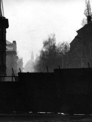Crno-bijela fotografija puste i maglovite sarajevske ulice pod snijegom, gdje se u prvom planu nalaze improvizovane metalne barikade za zaštitu građana od snajpera, dok se u pozadini naziru siluete zgrada i drveća.