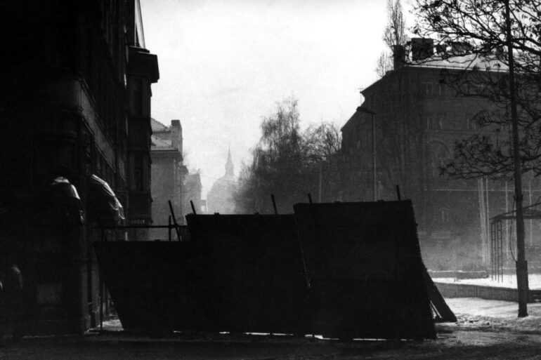Crno-bijela fotografija puste i maglovite sarajevske ulice pod snijegom, gdje se u prvom planu nalaze improvizovane metalne barikade za zaštitu građana od snajpera, dok se u pozadini naziru siluete zgrada i drveća.