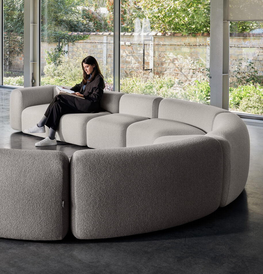 Volumen Dolmen modularna sofa u sivoj bouclé tkanini zaobljeni segmenti polukružna kompozicija, žena čita knjige na sofi uz stakleni zid s pogledom na vrt