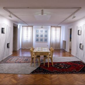 Panoramski pogled na centralnu salu postave Domus Diasporica — valovite linije na bočnim zidovima, orijentalni ćilimi na parketu, drveni sto u sredini i crteži u pozadini, Palazzo Malipiero, Venecija 2026.