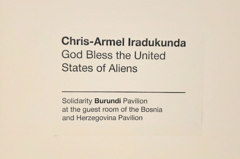 Zidna natpisna pločica s imenom umjetnika i nazivom rada: Chris-Armel Iradukunda, God Bless the United States of Aliens — Solidarity Burundi Pavilion at the guest room of the Bosnia and Herzegovina Pavilion, Palazzo Malipiero, Venecija 2026.