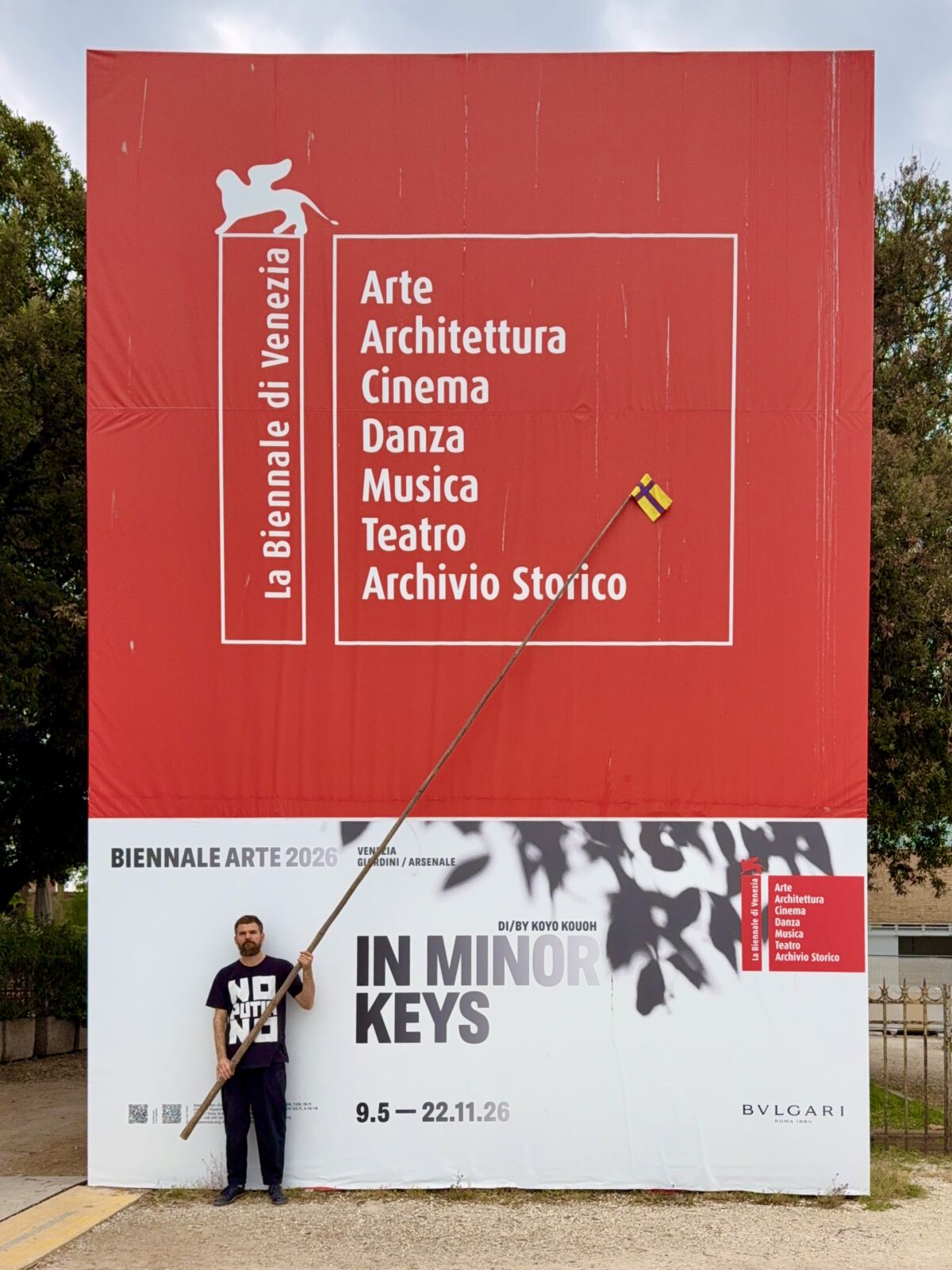 Pavel Rotts stoji pred velikim plakatom Biennale Arte 2026 s temom In Minor Keys kustosa Koyo Kouoha, držeći dugačku šipku s malom ingrijanskom zastavom na vrhu.