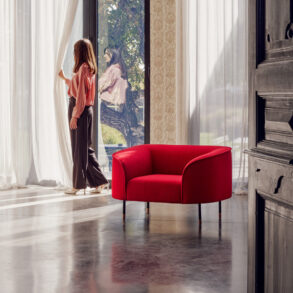 Volumen Le Mans fotelja crvena tkanina zaobljena forma metalne noge bakreni detalj, žena stoji pored prozora s bijelim zavjesama i refleksijom u staklu
