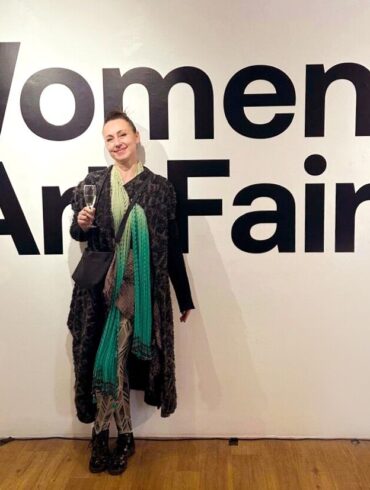 Aleksandra Nina Knežević, grafička dizajnerica i ilustratorica iz Sarajeva, na otvorenju Women in Art Fair CIC u Londonu 2026. — stoji ispred velikog natpisa izložbe, u crnom kaputu s tirkiznim šalom, s čašom šampanjca u ruci