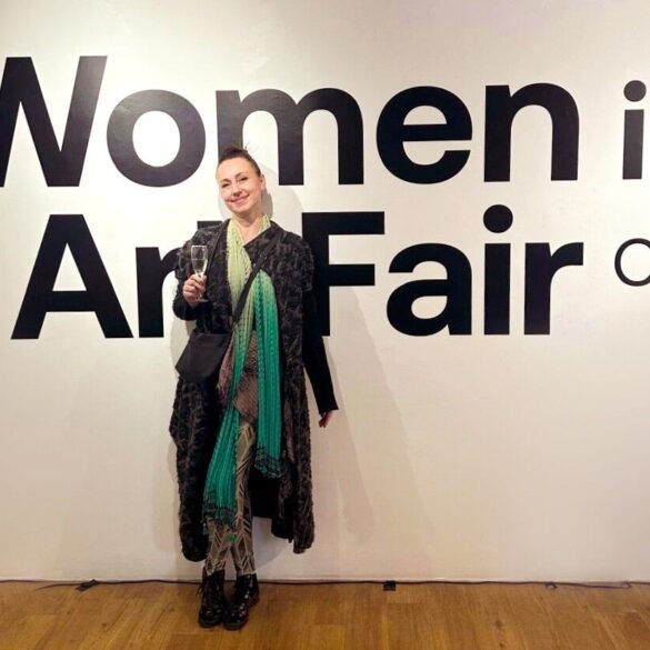 Aleksandra Nina Knežević, grafička dizajnerica i ilustratorica iz Sarajeva, na otvorenju Women in Art Fair CIC u Londonu 2026. — stoji ispred velikog natpisa izložbe, u crnom kaputu s tirkiznim šalom, s čašom šampanjca u ruci