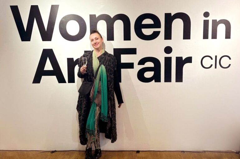 Aleksandra Nina Knežević, grafička dizajnerica i ilustratorica iz Sarajeva, na otvorenju Women in Art Fair CIC u Londonu 2026. — stoji ispred velikog natpisa izložbe, u crnom kaputu s tirkiznim šalom, s čašom šampanjca u ruci