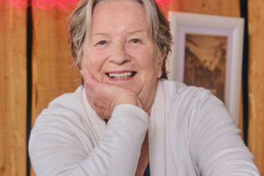 Portret nasmijane Sandy Myhre, osnivačice organizacije Women’s Worldwide Car of the Year (WWCOTY) i autorice knjige „One Lady Owner“, u bijelom džemperu sa naslonjenom rukom na bradu.