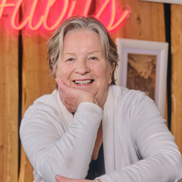 Portret nasmijane Sandy Myhre, osnivačice organizacije Women’s Worldwide Car of the Year (WWCOTY) i autorice knjige „One Lady Owner“, u bijelom džemperu sa naslonjenom rukom na bradu.