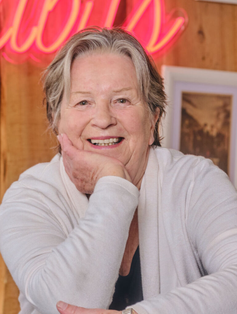 Portret nasmijane Sandy Myhre, osnivačice organizacije Women’s Worldwide Car of the Year (WWCOTY) i autorice knjige „One Lady Owner“, u bijelom džemperu sa naslonjenom rukom na bradu.