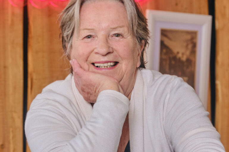 Portret nasmijane Sandy Myhre, osnivačice organizacije Women’s Worldwide Car of the Year (WWCOTY) i autorice knjige „One Lady Owner“, u bijelom džemperu sa naslonjenom rukom na bradu.