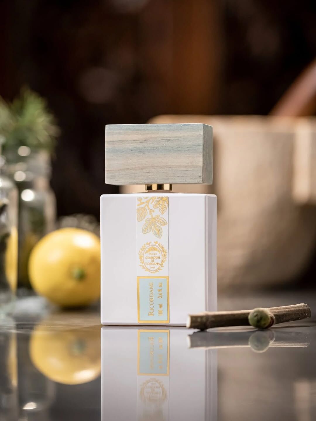 Giardini di Toscana Ricordami parfem 100ml u bijeloj bočici s blijedo plavim čepom i zlatnim listovima smokve, uz žuti citrus i grančicu u pozadini na sjajnoj podlozi