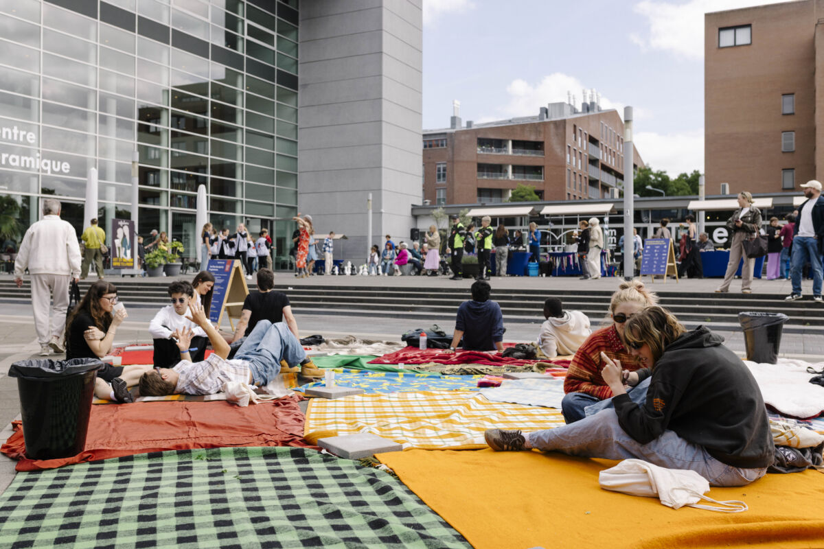 Posjetioci sjede i leže na šarenim ćebadima ispred zgrade Centre Céramique na Plein 1992, opušteni na zajedničkom pikniku tokom TexTiles BlockParty, u pozadini aktivnosti i nastup na trgu, Maastricht 2026.