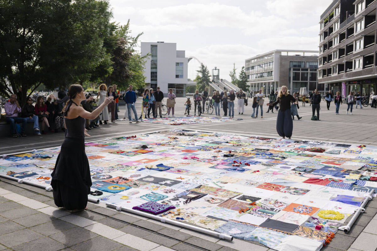 Dvije izvođačice u crnoj odjeći kleče i stoje s obje strane monumentalnog tekstilnog platna TexTiles položenog na Plein 1992, dok publika promatra performans u pozadini, Maastricht 2026.