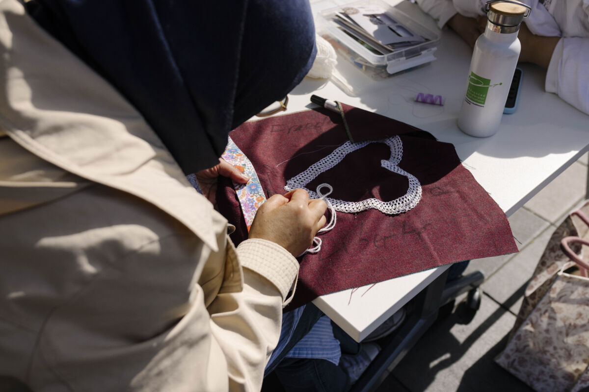Ruke žene s hidžabom koje šiju bijelu čipku u obliku srca na tamnocrvenoj tkanini za TexTiles pločicu, uz pribor za šivanje i bocu vode na stolu, radionica na otvorenom, Maastricht 2026.