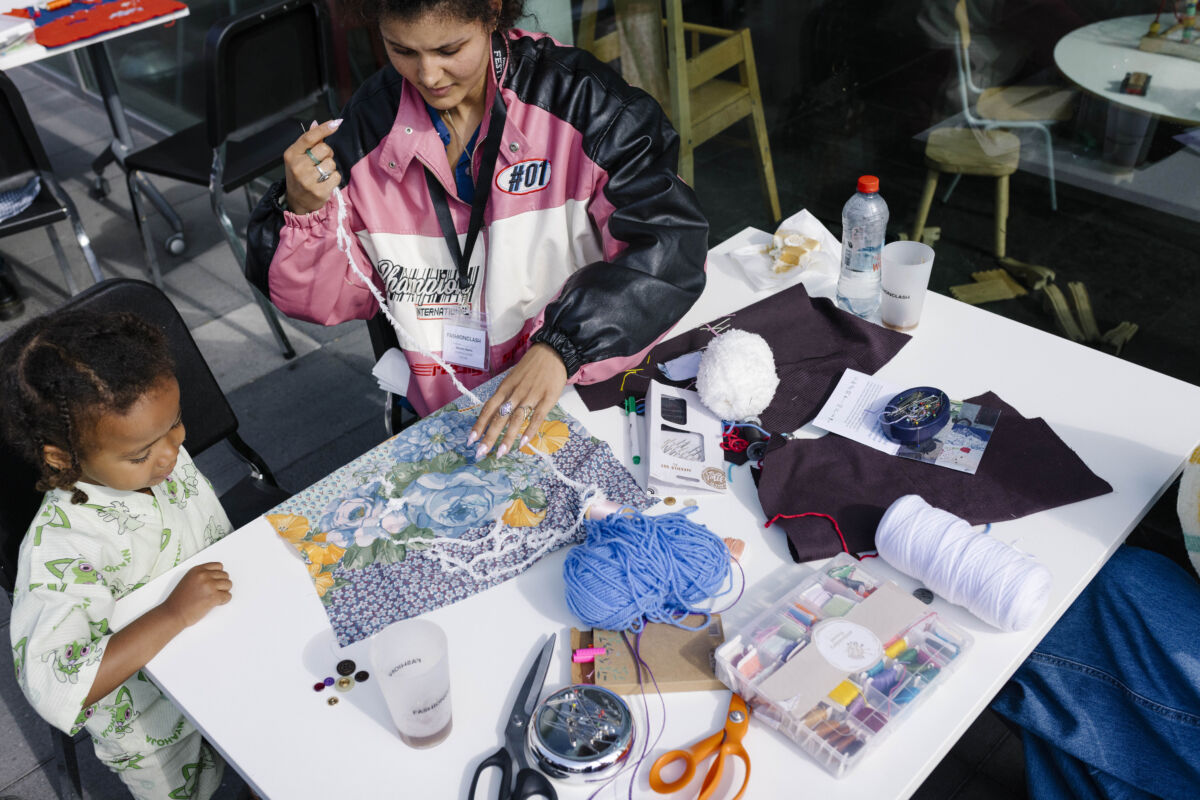 Žena u roze-crnoj jakni s natpisom FASHIONCLASH šije plocicu s cvjetnim motivom za TexTiles, dok mala curka u bijeloj majici sjedi pored nje i posmatra, okružene priborom za šivanje, Maastricht 2026.