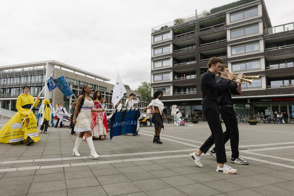 Svečana povorka izvođača u avangardnim kostimima prolazi Plein 1992 noseći zastave i transparent s natpisom FASHIONCLASH, dok trubači sviraju u prvom planu, otvaranje TexTiles BlockParty, Maastricht 2026.