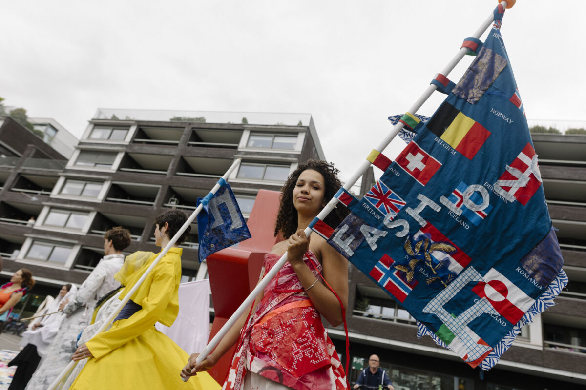 Mlada žena u crvenoj haljini s tradicijskim vezom nosi veliku plavu zastavu s izvezenim zastavama europskih zemalja i natpisom "FASHIONCLASH" tokom otvaranja TexTiles BlockParty na Plein 1992, Maastricht 2026.