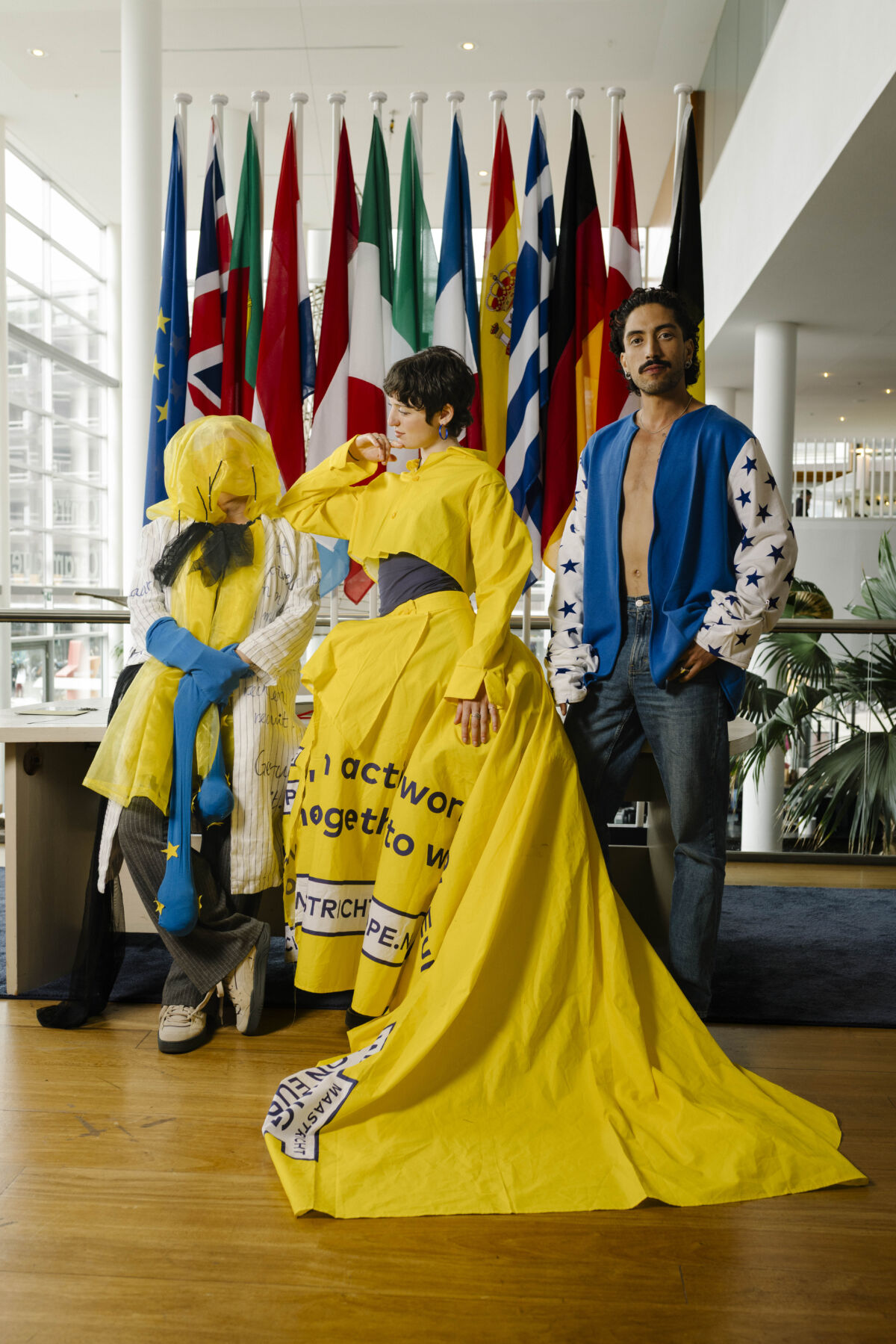 Trojica izvođača u avangardnim kostimima inspiriranim Evropom — žuta haljina s natpisima i plava jakna sa zvjezdicama — poziraju ispred zastava europskih zemalja za performans EUROPE BUT FASHION, Maastricht 2026.