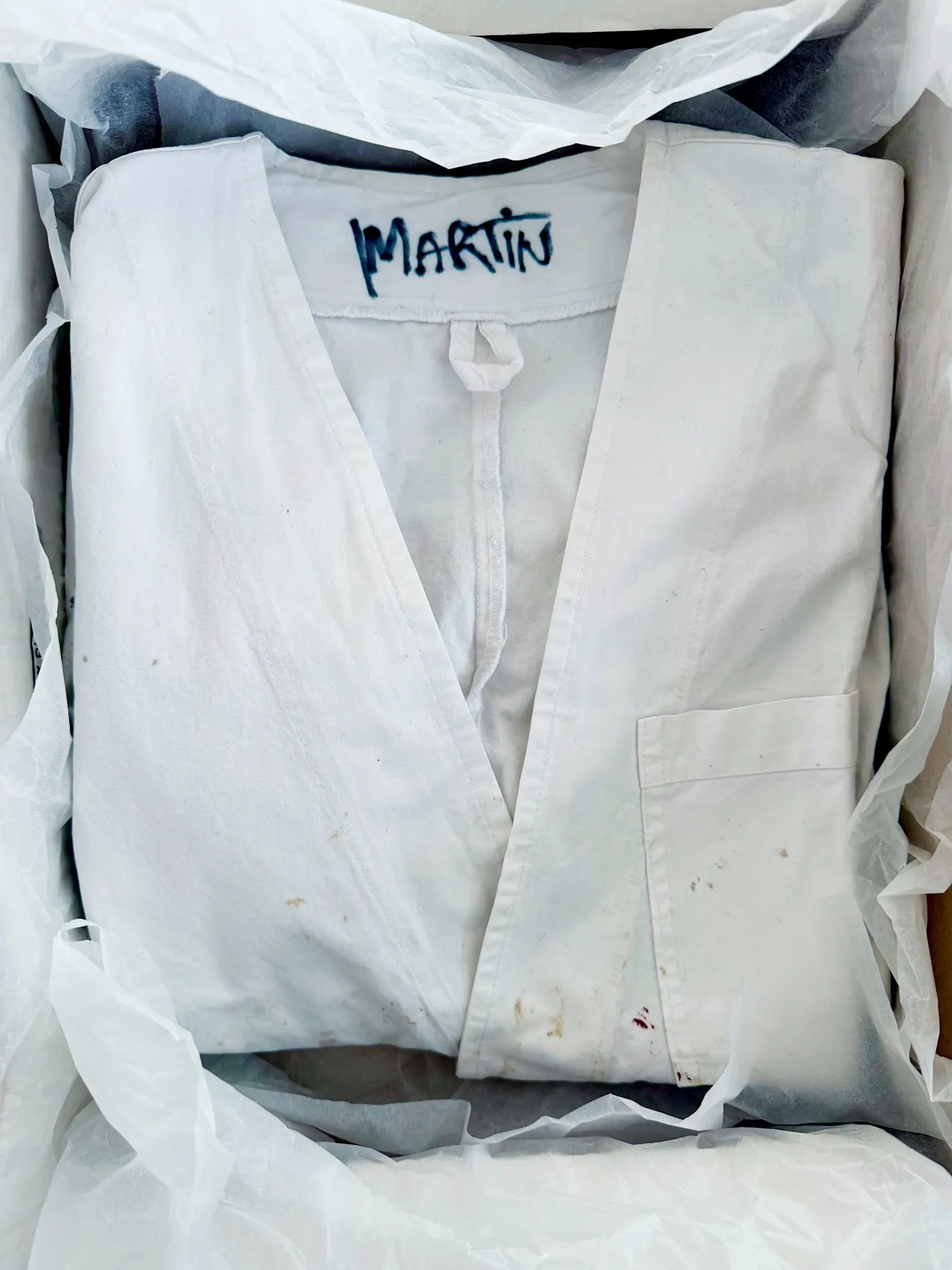 Bijela radna pregača Blouse Blanche s imenom Martin ispisanim flomasterom na ovratniku, složena u kutiji s bijelim papirom