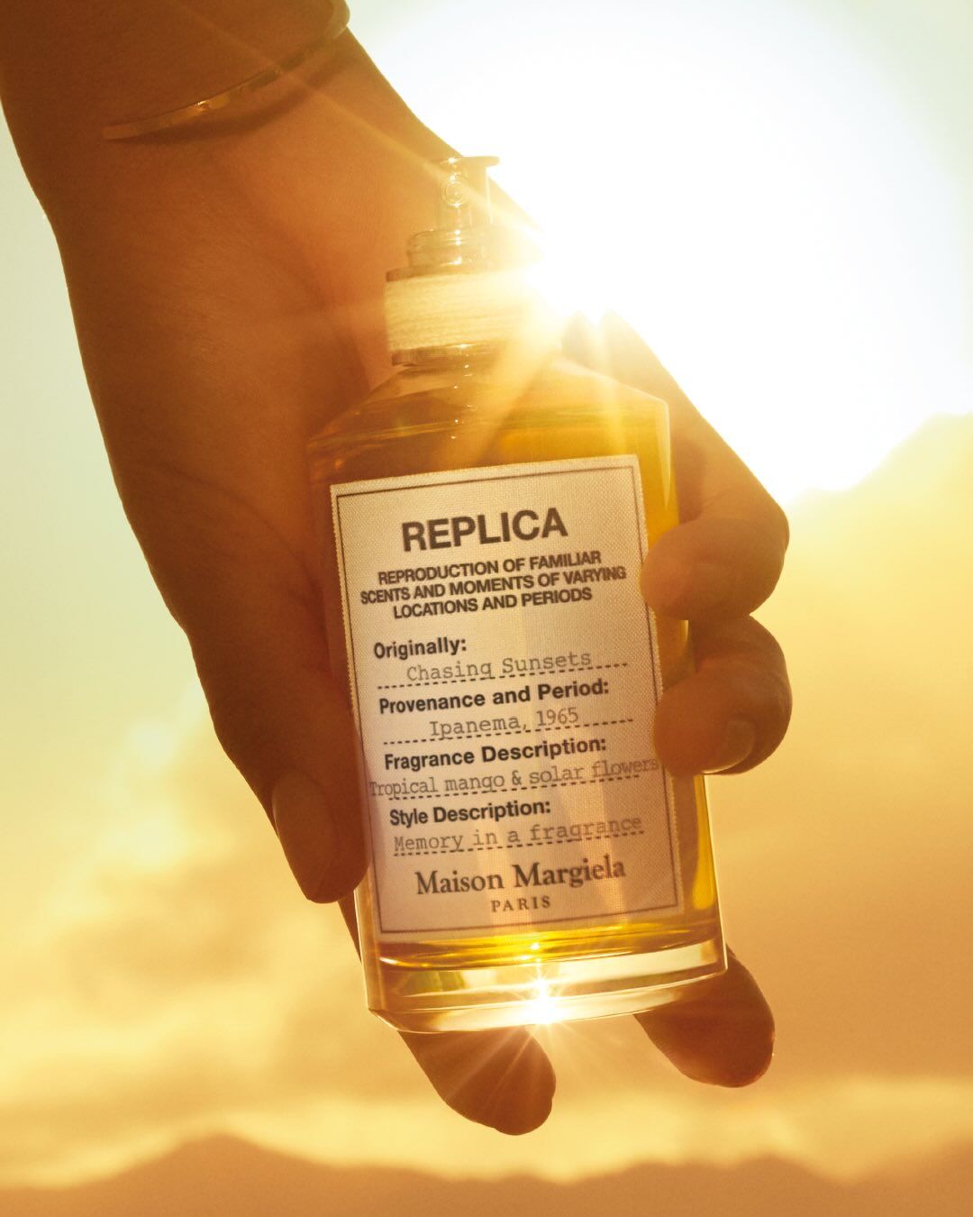 Ruka drži bočicu Maison Margiela Replica Chasing Sunsets prema suncu