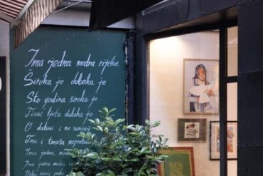 Vanjski izgled galerije Mezzanin na Ferhadiji 16 u Sarajevu, sa zelenom tablom ispisanom stihovima i izloženim radovima u izlogu