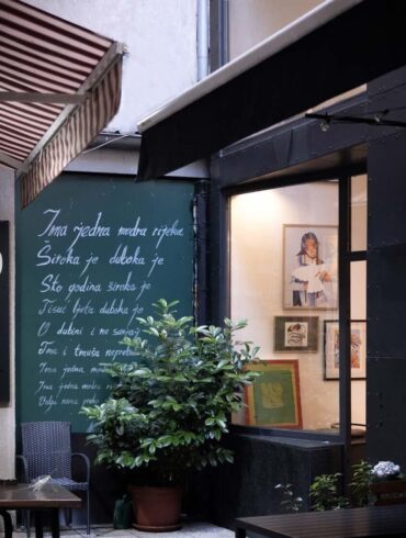 Vanjski izgled galerije Mezzanin na Ferhadiji 16 u Sarajevu, sa zelenom tablom ispisanom stihovima i izloženim radovima u izlogu