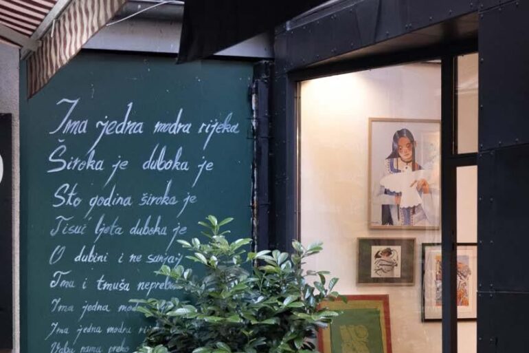 Vanjski izgled galerije Mezzanin na Ferhadiji 16 u Sarajevu, sa zelenom tablom ispisanom stihovima i izloženim radovima u izlogu
