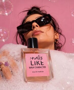 Martimex parfimerija - žena drži bočicu Smells Like Main Character parfema okružena mehurićima na roza pozadini