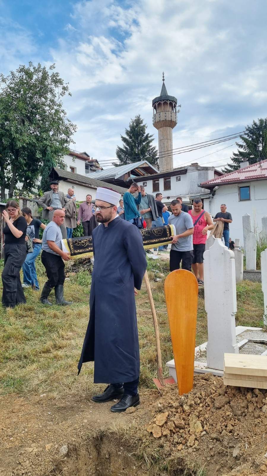 Hodža u tradicionalnoj odori stoji pored otvorenog groba tokom snimanja scene ukopa, u pozadini minaret džamije