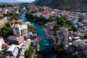 Ptičja perspektiva Starog mosta i rijeke Neretve tokom Red Bull Cliff Diving takmičenja u Mostaru, s dva paraglajdera iznad vode i mnoštvom posjetitelja na obali
