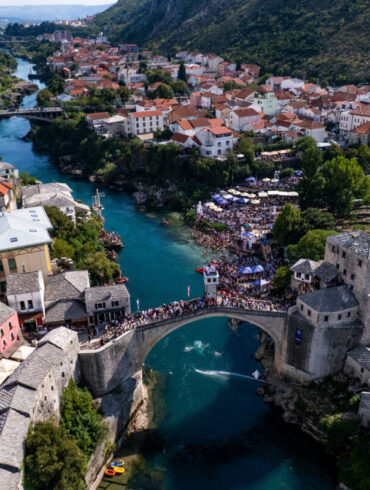 Ptičja perspektiva Starog mosta i rijeke Neretve tokom Red Bull Cliff Diving takmičenja u Mostaru, s dva paraglajdera iznad vode i mnoštvom posjetitelja na obali