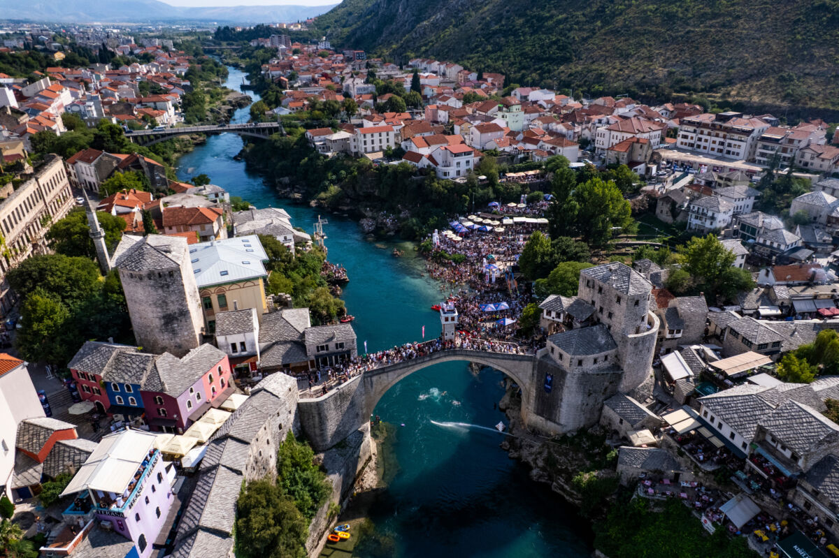 Ptičja perspektiva Starog mosta i rijeke Neretve tokom Red Bull Cliff Diving takmičenja u Mostaru, s dva paraglajdera iznad vode i mnoštvom posjetitelja na obali