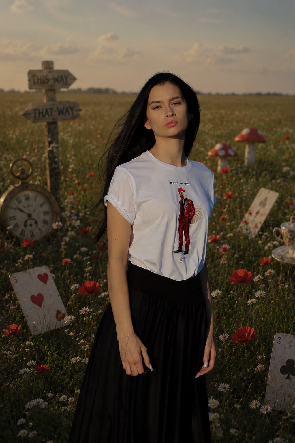 Model u bijeloj majici s printom i crnoj suknji stoji u polju divljeg cvijeća pored drvenog putokaza "This Way / That Way" – kampanja "Alisa u Bosni". Foto i AI postprodukcija: HAAD produkcija