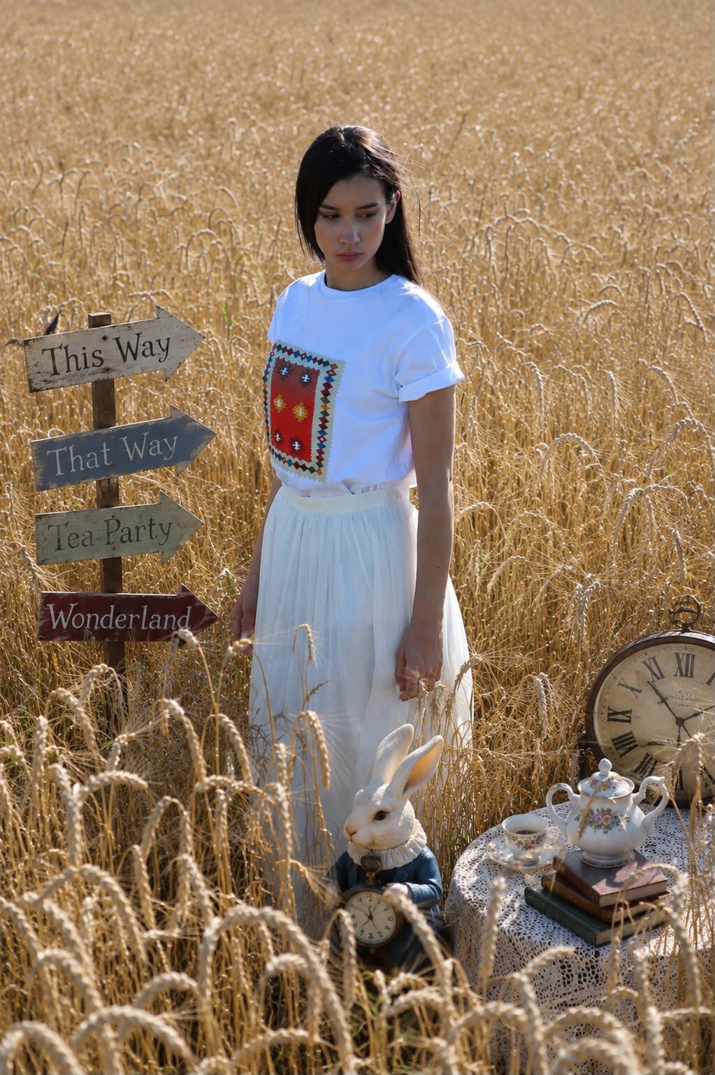 Model u bijeloj majici s bosanskim motivom i bijeloj suknji stoji u žitnom polju pored putokaza sa natpisima "This Way, That Way, Tea Party, Wonderland" – HAAD modna priča. Foto i AI postprodukcija: HAAD produkcija