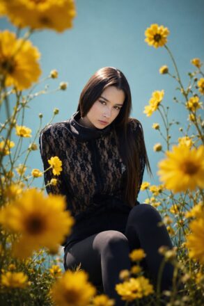 Model čučne u polju žutog cvijeća, odjevena u crnu čipkastu jaknu i tajice – urbani stil HAAD kolekcije "Alisa u Bosni". Foto i AI postprodukcija: HAAD produkcija