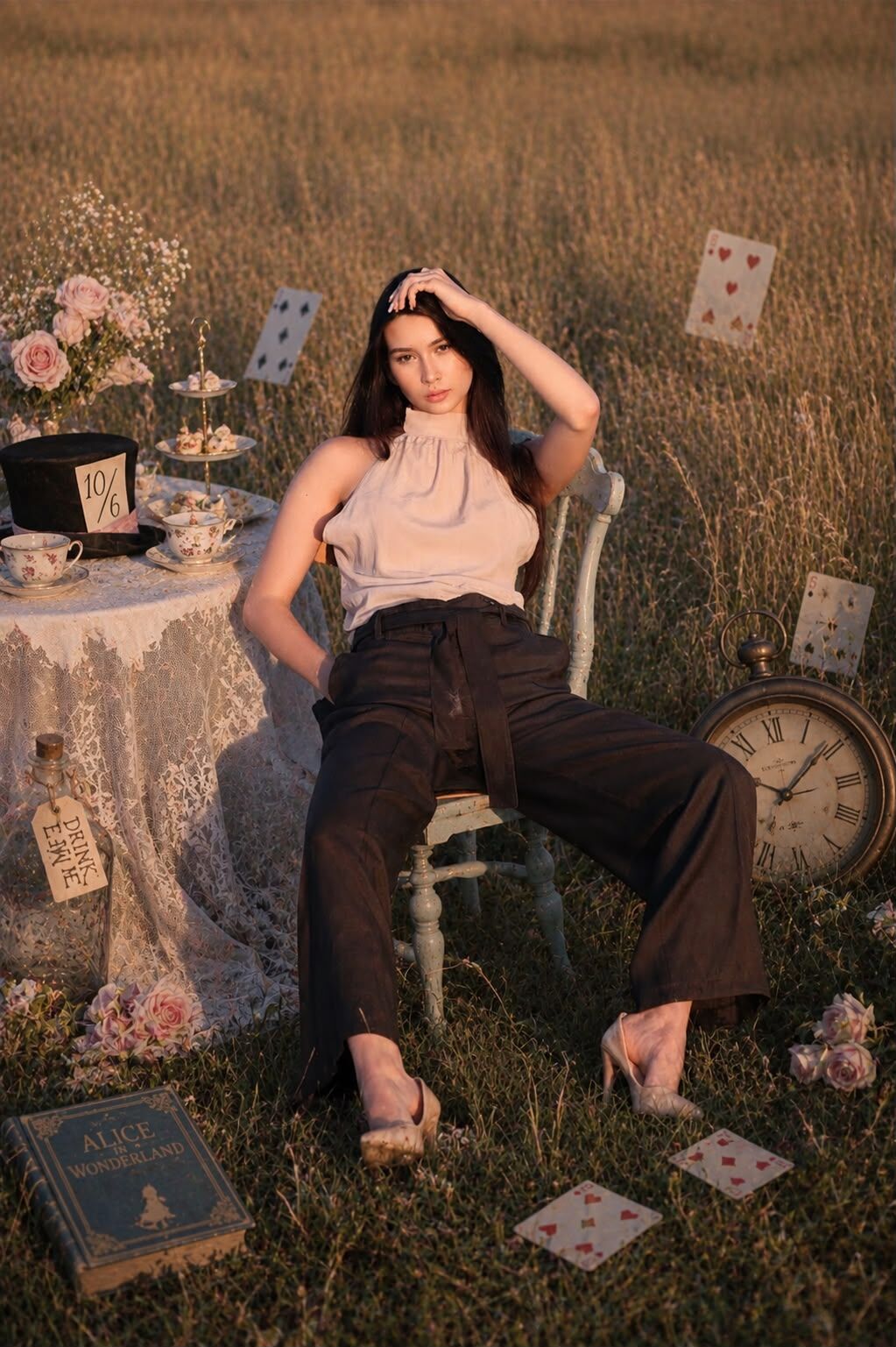 Model u bež bluzi i tamnosmeđim pantalonama sjedi na stolici pored čajnog stola u polju, s knjigom "Alice in Wonderland" u prvom planu – HAAD kampanja snimljena u Bosanskom Petrovcu. Foto i AI postprodukcija: HAAD produkcija