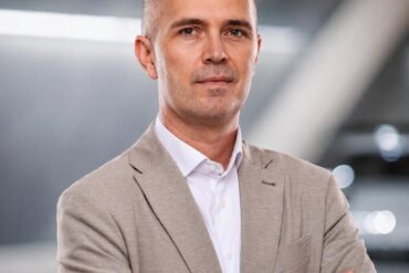 Portret Ismira Omeragića, Brand Managera Škode za Bosnu i Hercegovinu, u bež blazeru i beloj košulji sa automobilskom infrastrukturom u blure pozadini