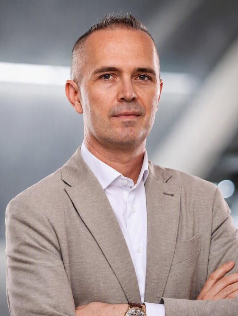 Portret Ismira Omeragića, Brand Managera Škode za Bosnu i Hercegovinu, u bež blazeru i beloj košulji sa automobilskom infrastrukturom u blure pozadini