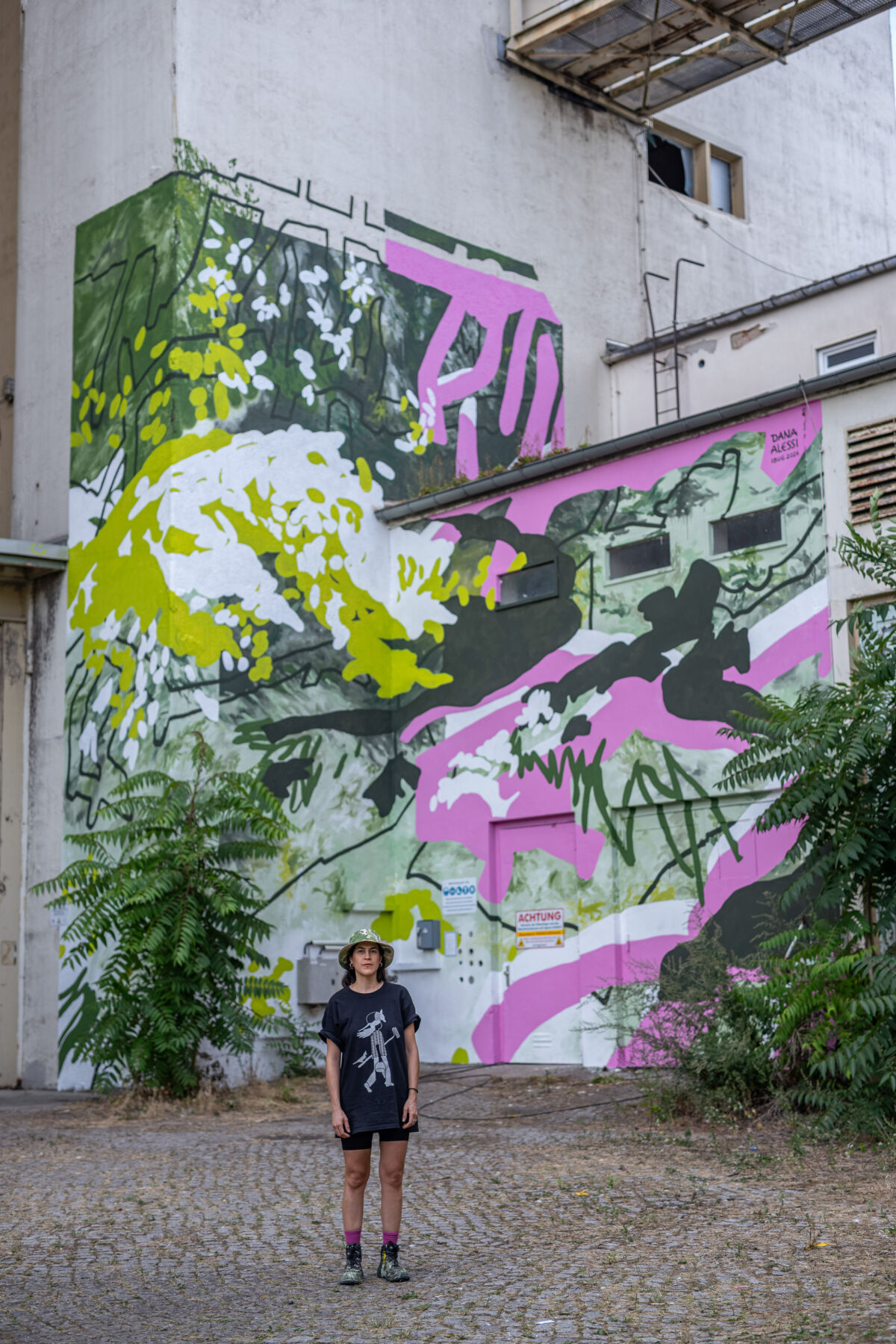 Veliki mural Dane Alessi na industrijskoj zgradi na IBUG Festivalu 2026., u zelenim, ružičastim, bijelim i crnim tonovima.