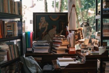 Kućna biblioteka s policama punim knjiga i naslaganim nepročitanim naslovima na radnom stolu